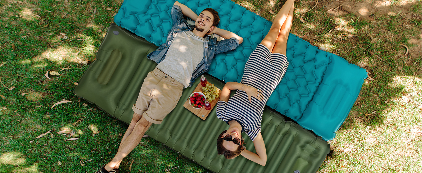 Two people relaxing on inflatable camping mattresses outdoors. Green and teal mattresses on grass with people lying down and enjoying snacks.