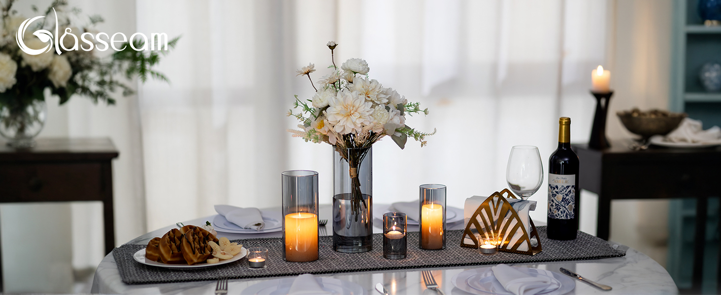 Elegante Tischdekoration mit Weinflaschen, Vasen mit weißen Blumen, Orangensaftgläsern und etwas, das aussieht wie Gebäck oder Brot auf einer langen dunklen Oberfläche vor weißen Vorhängen.