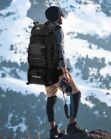 Personne portant un grand sac à dos noir debout sur un éperon rocheux, surplombant un paysage de montagnes enneigées. Démontre l'utilisation de l'équipement de plein air dans des conditions hivernales.