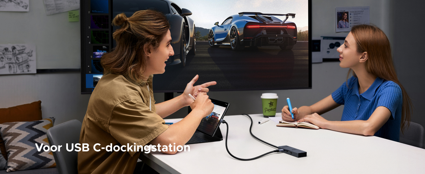 Zwei Leute diskutieren an einem Schreibtisch, hinter ihnen ein großer Bildschirm, auf dem Sportwagen zu sehen sind. Eine USB-C-Dockingstation ist auf dem Tisch sichtbar