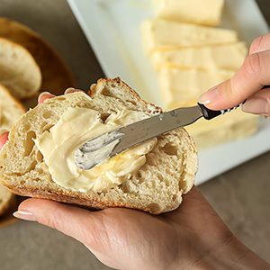 Primer plano de una mano untando mantequilla sobre una rebanada de pan con un cuchillo. Barra de pan parcialmente visible en segundo plano