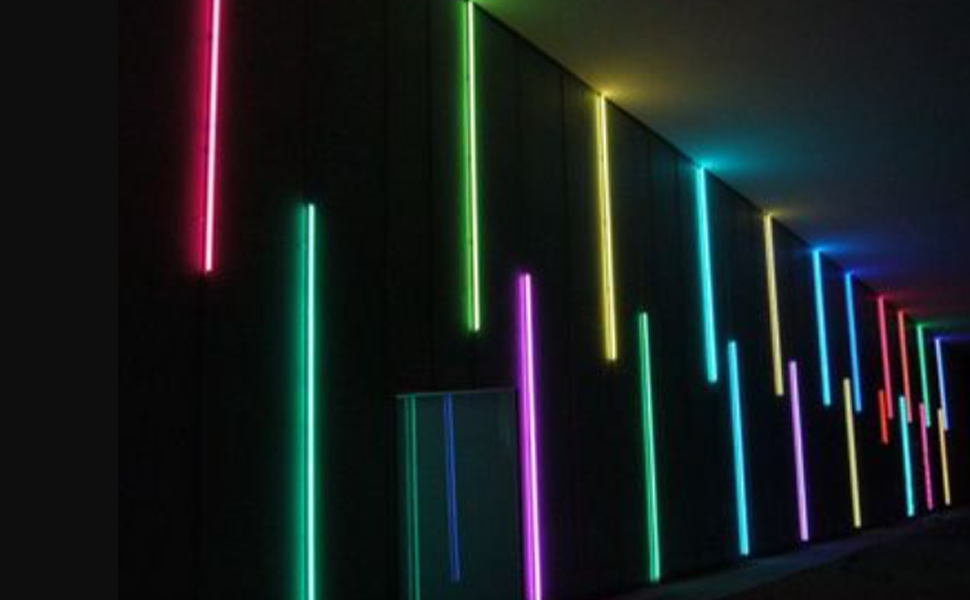 Vertical light tubes in various colors (pink, green, yellow, blue) arranged in a row along a dark wall or ceiling.