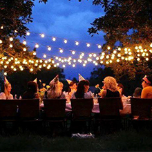 Outdoor evening gathering with string lights overhead. People seated at a long table in a garden or backyard setting, creating a festive atmosphere.