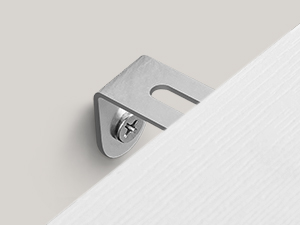 Közelkép egy kis, ezüstszínű fém rögzítőelemről vagy kampóról, amely fehér felületre van szerelve. A rögzítőelem a dőlésgátló rendszer része.