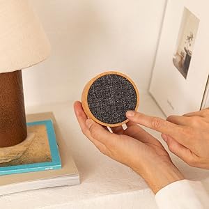 Manos que sostienen un pequeño dispositivo circular con una superficie de textura oscura, posiblemente un altavoz o un mando doméstico inteligente, junto a una lámpara sobre una mesa