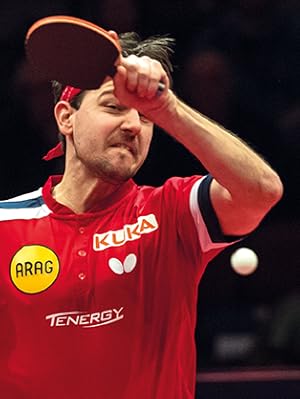 Jugadora de tenis de mesa en acción vistiendo una camiseta deportiva roja con los logotipos de los patrocinadores visibles, y se muestra ejecutando una técnica de juego durante un partido.