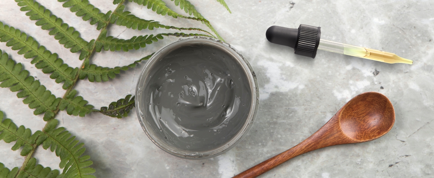 jar of gray clay mask with a dropper and wooden spoon on a marble surface, flanked green fern leaves