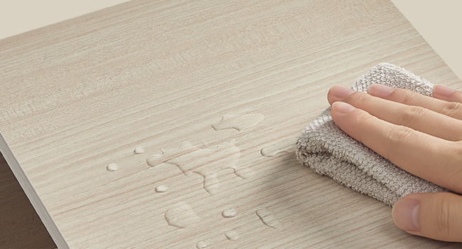 Kvapky vody na svetlom drevenom povrchu sa zotriú sivou handričkou a vykazujú vodeodolné alebo vodotesné vlastnosti.