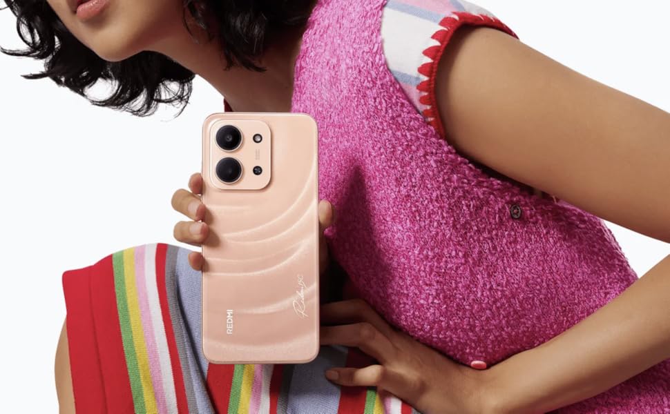 Close-up of hands holding a peach-colored smartphone, wearing pink textured top. Rainbow-striped fabric visible in background.