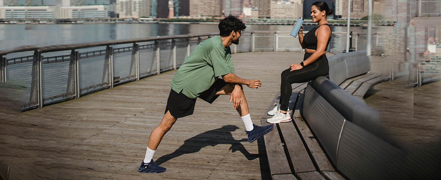 Two people exercising on an urban waterfront. One stretches their leg on a bench while the other observes. Both wear athletic clothing and sneakers.