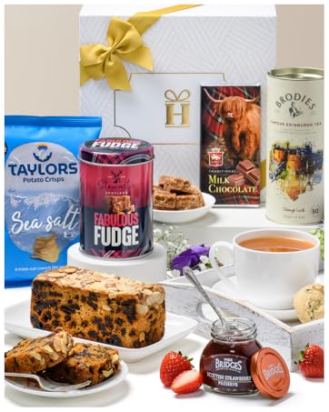 Tea and cake gift set featuring fruit cake slices, strawberry jam, sea salt snacks, fudge, and chocolate products arranged with teacups and decorative bows. Various branded packages visible with 'Taylors' tea clearly shown.