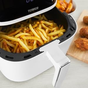 White air fryer with black basket containing golden french fries, shown in use with fried chicken pieces on wooden board nearby.