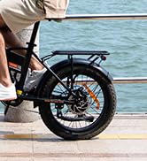 Vista parcial de la rueda trasera de una bicicleta eléctrica con neumático grueso, freno de disco y cremallera trasera, colocada cerca del agua.