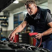 Mechanic working on car engine, holding red diagnostic tool. Wearing dark clothing in dimly lit garage or workshop setting.