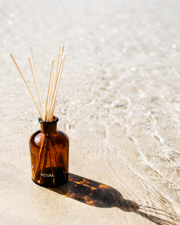 Amber glass bottle with reed diffuser sticks on sandy beach. Bottle labeled 'HEVAI'. Shallow water visible, creating reflective background.
