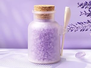 Glasgefäß mit Korkdeckel mit violettem Badesalz, dazu eine weiße Schaufel und Lavendelzweige vor