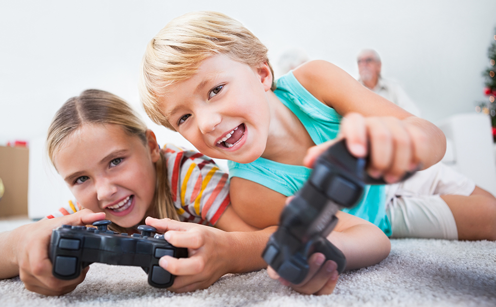 Dos niños tumbados en la alfombra, sonriendo mientras sostienen mandos negros de videojuegos. Parece que están jugando un juego juntos.