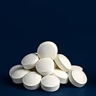 Diverse compresse o pillole farmaceutiche bianche e rotonde disposte su sfondo blu scuro. Le pillole appaiono uniformi per dimensioni e forma.