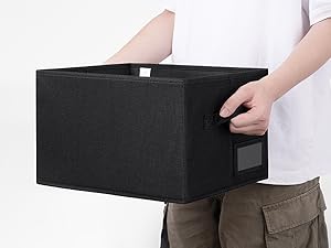 Person, die eine große schwarze Stoff-Aufbewahrungsbox mit Griffen in der Hand hält. Die Box scheint zusammenklappbar zu sein und hat auf einer Seite ein kleines Fenster oder einen Etikettenbereich