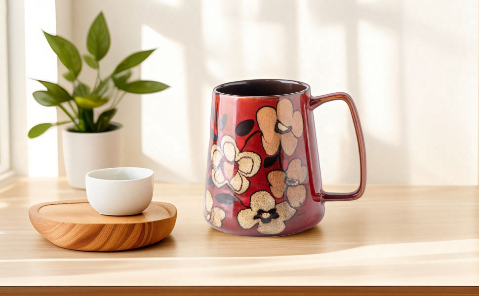 Red ceramic mug with floral pattern design, displayed on wooden surface next to small white bowl and green plant.