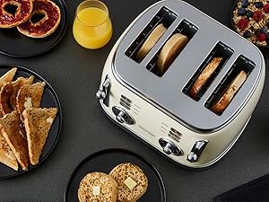 Stainless steel 4-slice toaster shown in use with toasted bread and orange juice on dark surface. Additional plates with sliced toast visible in scene.