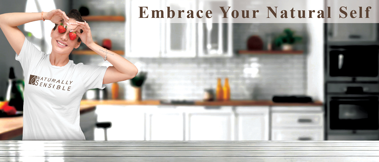Woman in a kitchen holding strawberries over her eyes, wearing a shirt that says Naturally Sensible