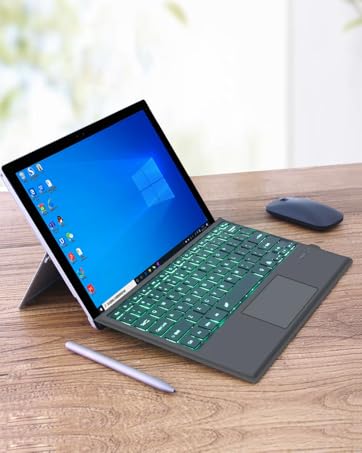 Tablet-style laptop with detachable keyboard on wooden desk. Features blue screen, backlit green keyboard, stylus, and wireless mouse. Device in tent mode, showing Windows interface.