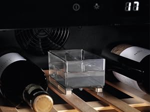 Clear acrylic display box positioned on wooden wine rack with dark wine bottles in background. Ambient lighting creates dramatic shadows.