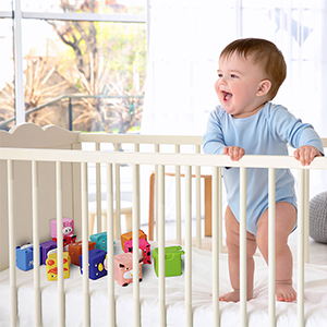 A baby is standing in a crib laughing happily, with some soft blocks on it