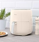 Cream-colored cylindrical air fryer on kitchen counter alongside decorative items and a plate of food. Features minimalist design with control panel.