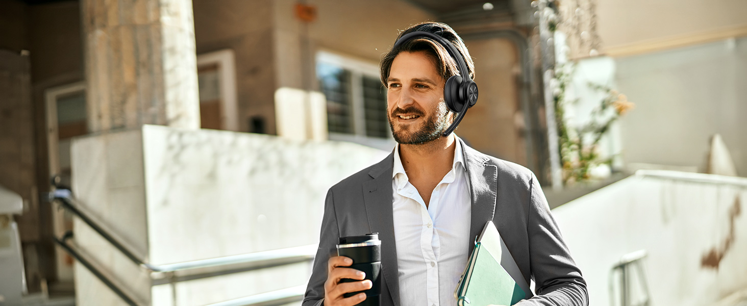Person wearing over-ear headphones and business attire holding a mobile device outdoors on a sunny terrace.