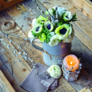 Stilllebenarrangement mit grünen Blumen in einer metallischen Vase und einer glühenden Kerze auf einer rustikalen Holzoberfläche.