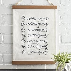 hanging canvas sign with be courageous lettering on a white brick wall above a shelf holding a plant