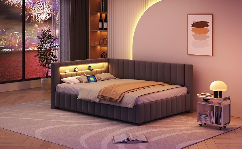 Modernes Schlafzimmer mit grauem Polsterbett, rosafarbenen Wänden und Umgebungsbeleuchtung. Verfügt über einen Nachttisch, Kunstwerke und einen Blick auf die Stadt mit einem Feuerwerk durch das Fenster