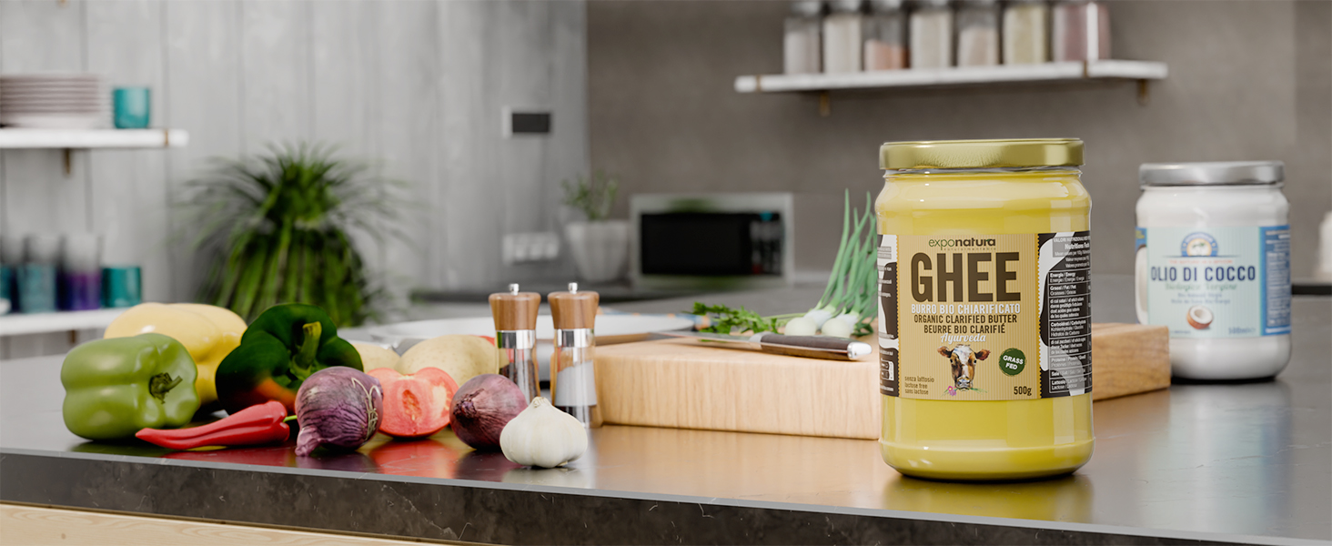 Bancone da cucina con vari ingredienti e un grande barattolo di burro chiarificato. Tagliere in legno, verdure fresche e vasetti più piccoli visibili sullo sfondo