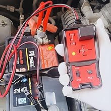 Car engine compartment with visible battery and red diagnostic tool connected. Tool has digital display and multiple connection points.