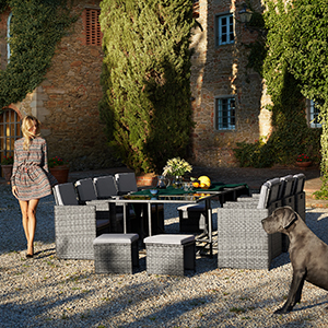avec chaises et table tissées. Femme en costume marchant à proximité. Grand chien au premier plan. Bâtiment en pierre et arbres en arrière-plan.