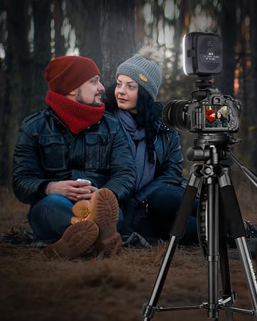 Camera on tripod with couple sitting nearby. Dark setting with people wearing winter clothing. Camera appears to be recording or photographing.