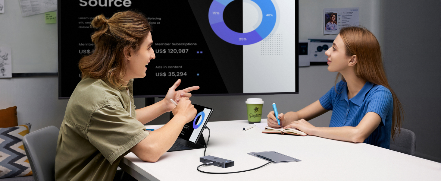 Two people sitting at a white table in an office setting. A large screen in the background displays a pie chart and text. One person is gesturing while the other takes notes.