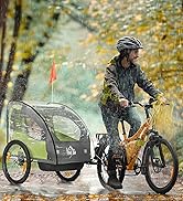 Cyclist riding an orange mountain bike in rain, towing a green and gray child trailer with safety flag. Autumn forest setting with falling leaves.
