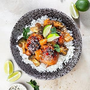 Asian-style dish with glazed chicken or protein served over white rice in a speckled black ceramic bowl, garnished with lime wedges and herbs.
