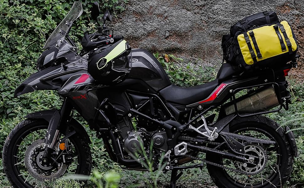 Black adventure motorcycle with windshield and side luggage, parked on dirt near rocky wall and vegetation. Features dual front lights and rugged off-road tires.