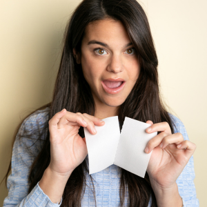 Mujer con camisa azul que sostiene dos pequeños objetos cuadrados blancos, posiblemente papeles o tarjetas, frente a ella con una expresión emocionada.