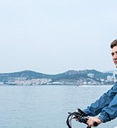 Vista panorámica del paseo marítimo con montañas distantes visibles a través de un cuerpo de agua, que muestra parcialmente el manillar de una bicicleta en primer plano