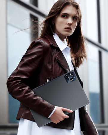 Professional setting showing figure in brown leather jacket and white shirt holding sleek gray laptop or device while standing against light-colored wall.