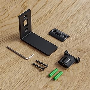 Components of a wall mounting kit including L-shaped bracket, screws, wall anchors, hex key, and mounting plates. All parts are black except for green wall anchors, displayed on a wooden surface.