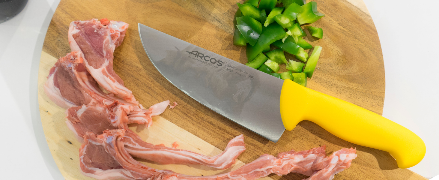 con mango amarillo sobre tabla de cortar de madera. Se ve carne cruda y pimientos verdes picados.