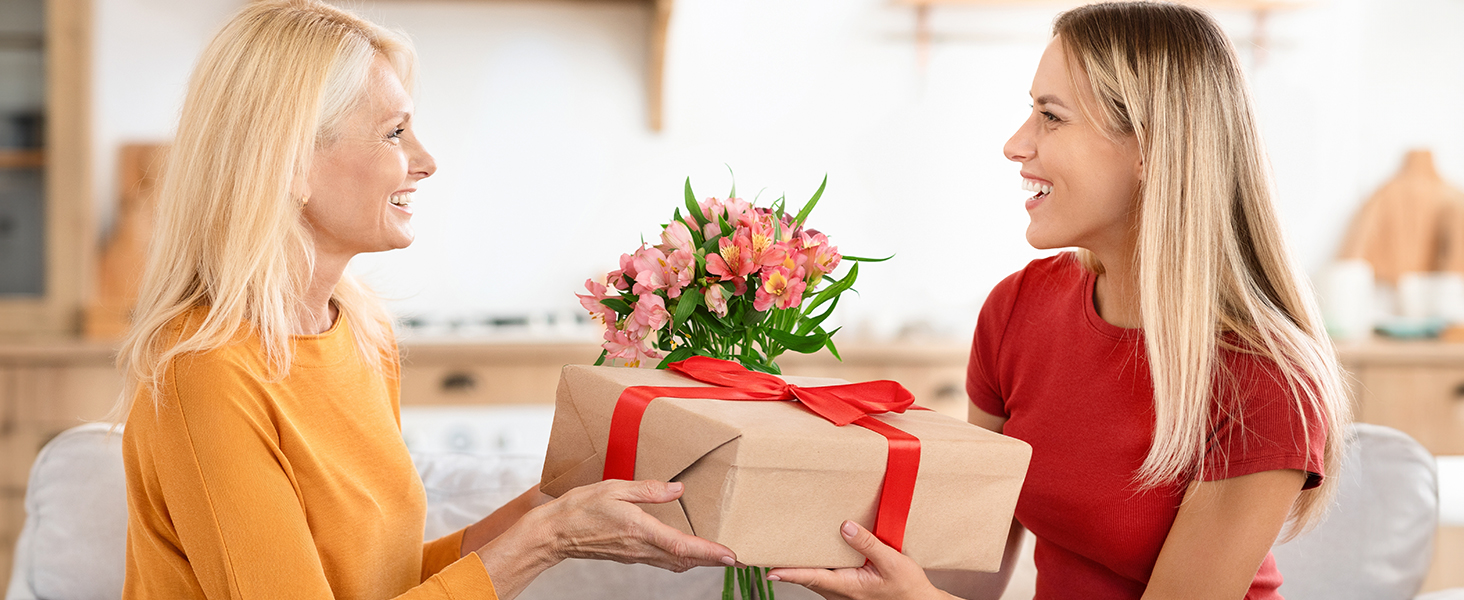 Two women in a kitchen, one handing the other a gift box with flowers. The box is wrapped in brown paper with a red ribbon.