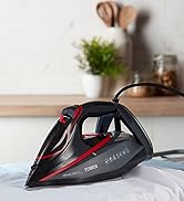 Modern black and red steam iron on white ironing surface with green plant in soft focus background.
