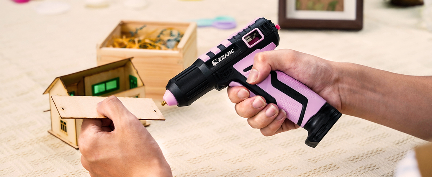 Black and pink cordless hot glue gun being used on a small wooden house model, with craft supplies visible in background.